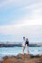 A romantic photo of a couple standing on a beach with crystal-clear blue water and a blue sky in the background. The man is holding the woman's hand as they look out towards the horizon